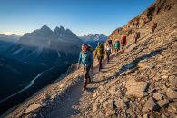 Hikers scramble along a steep rocky trail toward Ha Ling Peak with golden hour sunlight and panoramic mountain views in the background.