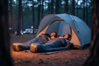 Family camping at dusk with sleeping bags near a dome tent in a wooded campsite, illuminated by warm campfire light.